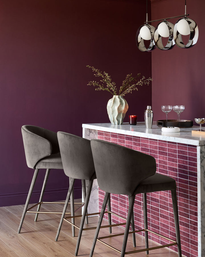 Ethan bar stool in dark chocolate velvet with tapered legs, shown at a modern bar with marble countertop and red brick panel.