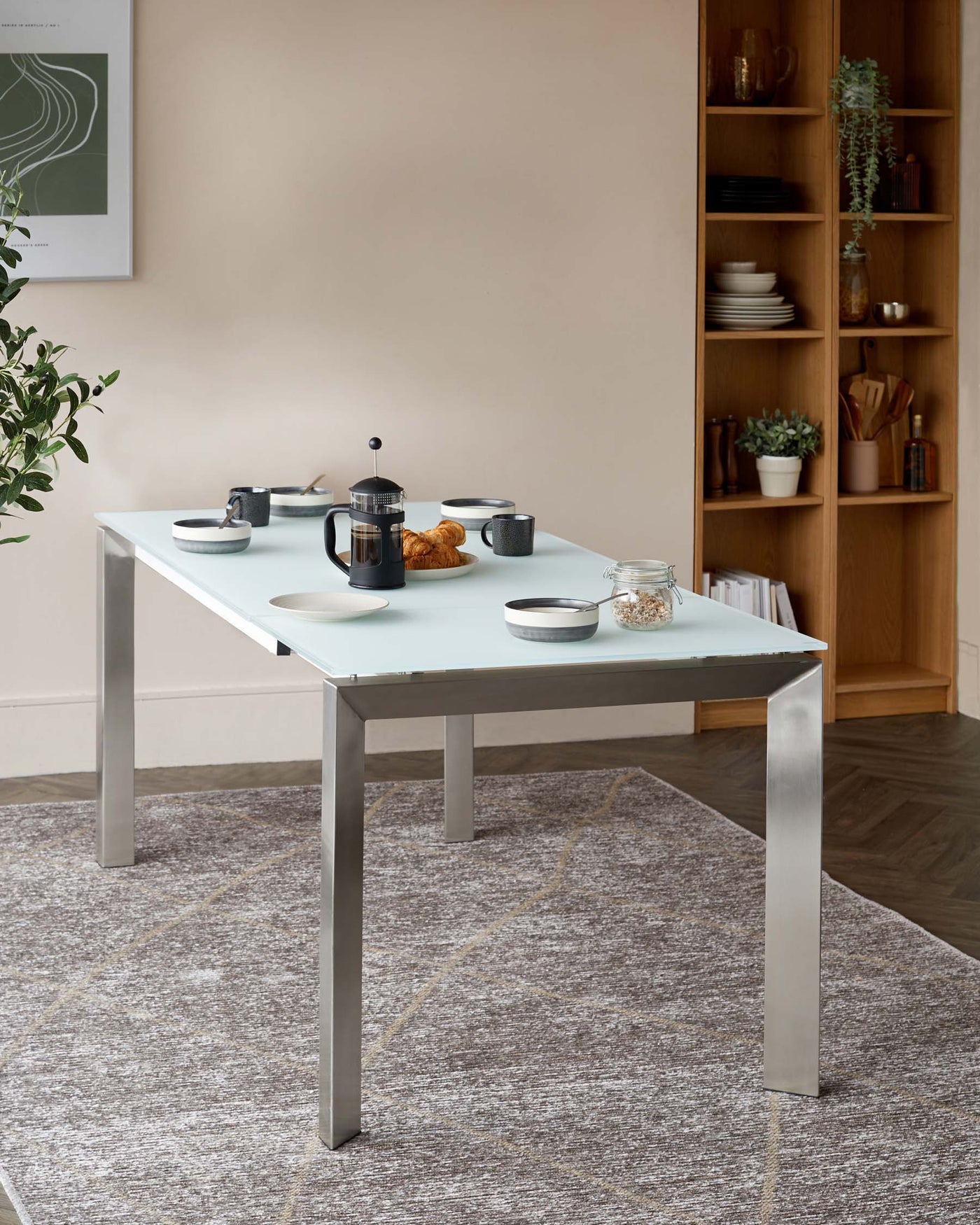 Modern minimalist dining table with a light grey tabletop and square stainless steel legs, accompanied by a tall wooden shelving unit with various decorative items and kitchenware.