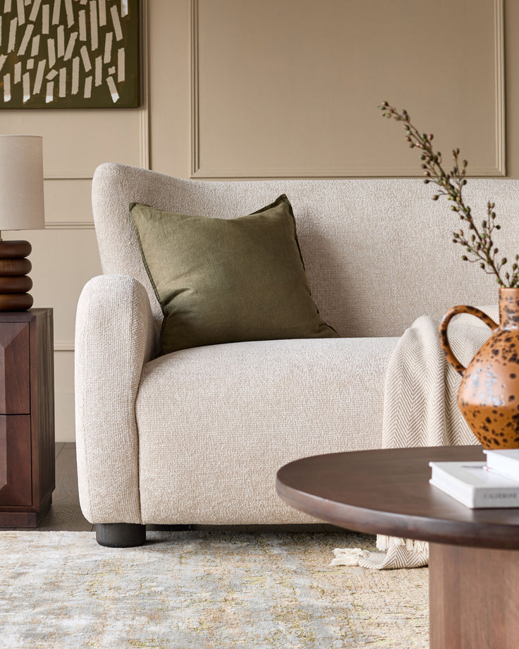 Evelyn 2-seater sofa in natural fabric with curved armrests, shown in a styled living room setting with olive pillow.