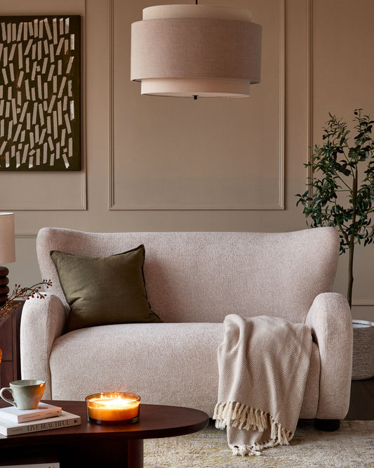 Evelyn 2-seater sofa in natural fabric, displayed in a warm living room setting with cushions and throw blanket.