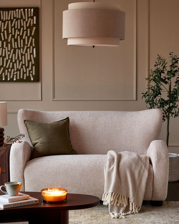 Evelyn 2-seater sofa in natural fabric, displayed in a warm living room setting with cushions and throw blanket.