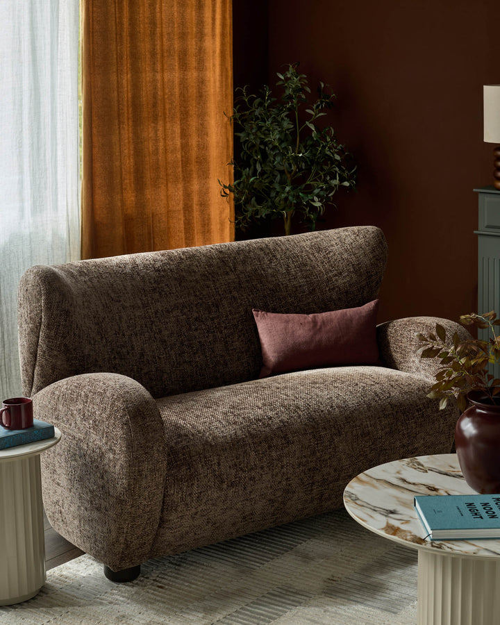 Evelyn 2-seater sofa in mocha fabric with curved armrests and high back, styled with burgundy pillow in warm interior setting.