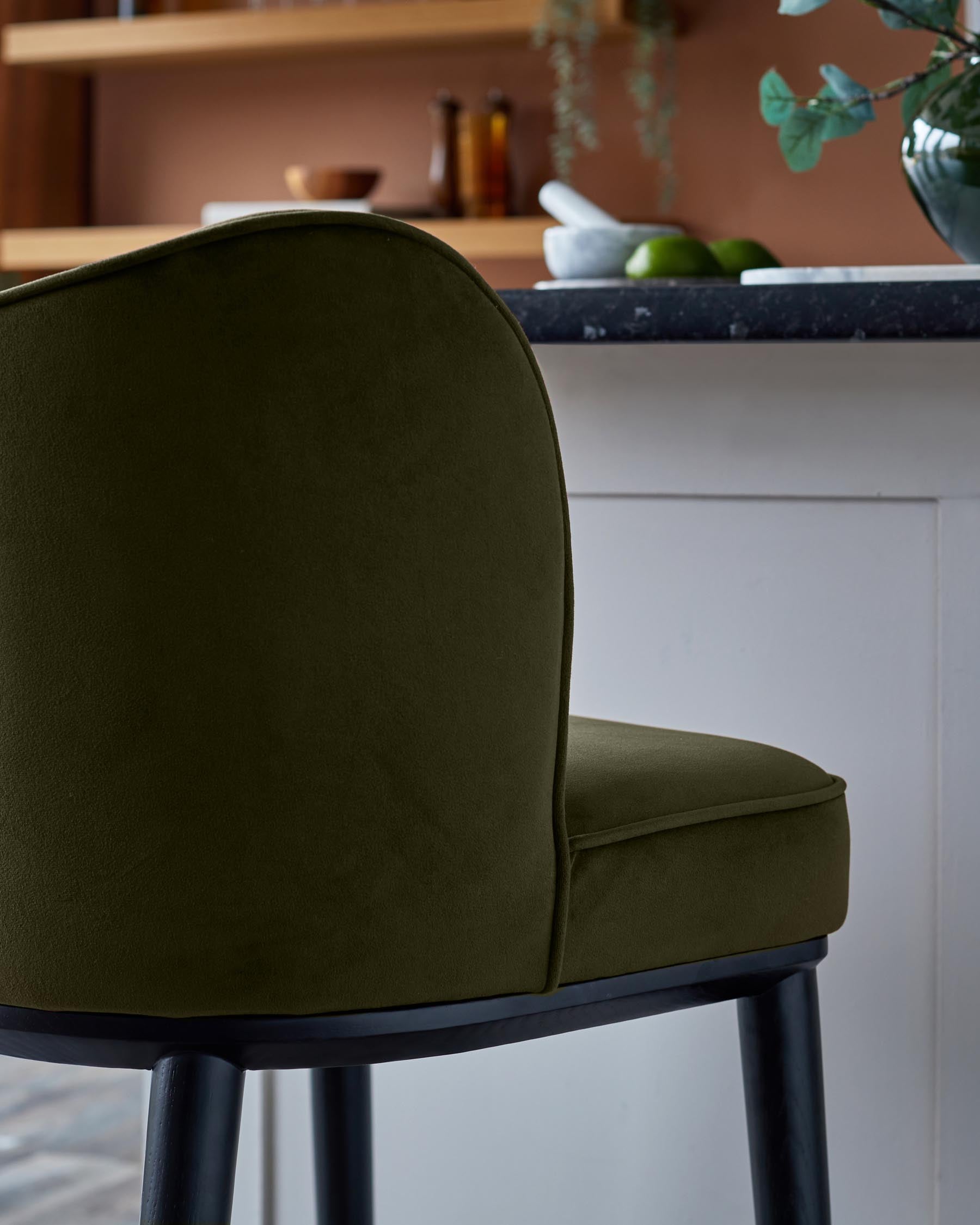 Close up of a dark green upholstered chair with black legs, set against a kitchen background.