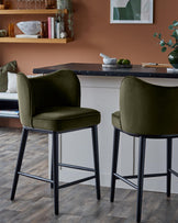 Two stylish olive green bar stools with black legs, sit under a kitchen counter in a modern kitchen.
