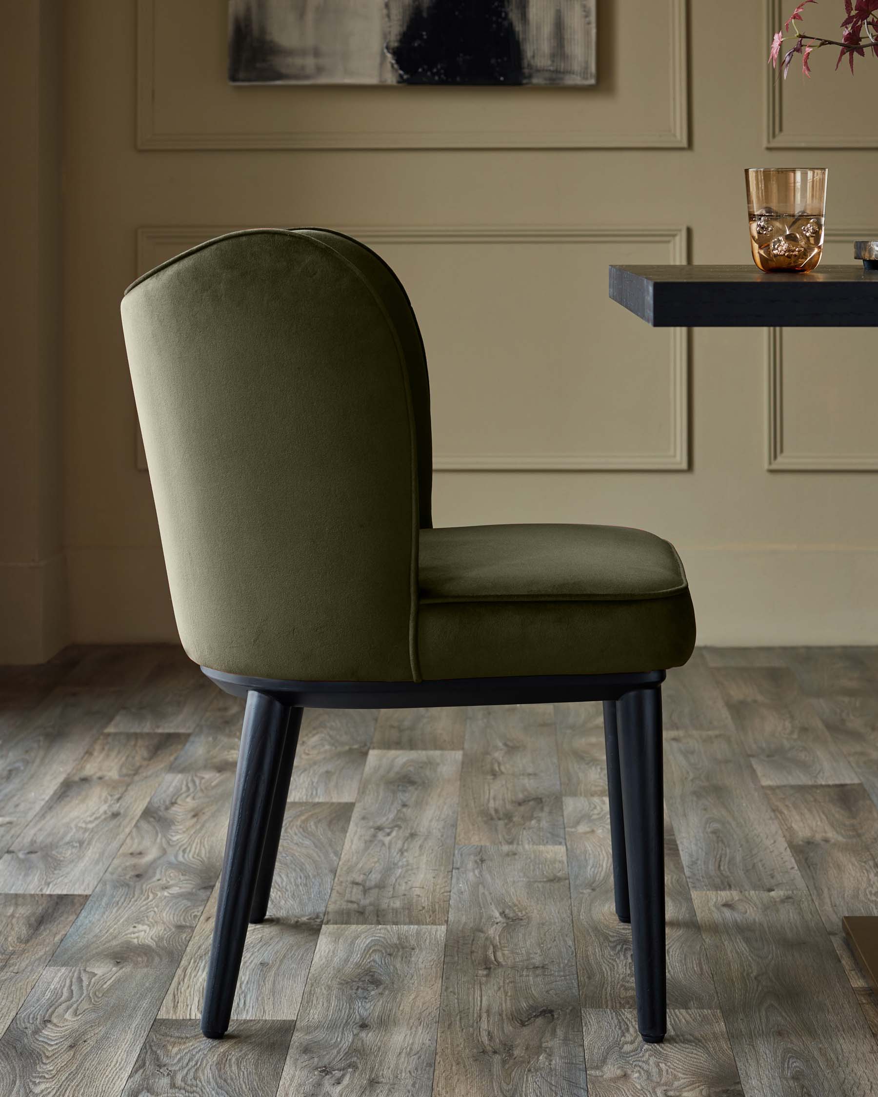 Olive green velvet dining chair with black legs, sat in front of a dark table.