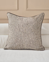 Fabian cushion in natural textured weave with dark piping, displayed on a cream upholstered sofa against a beige paneled wall.