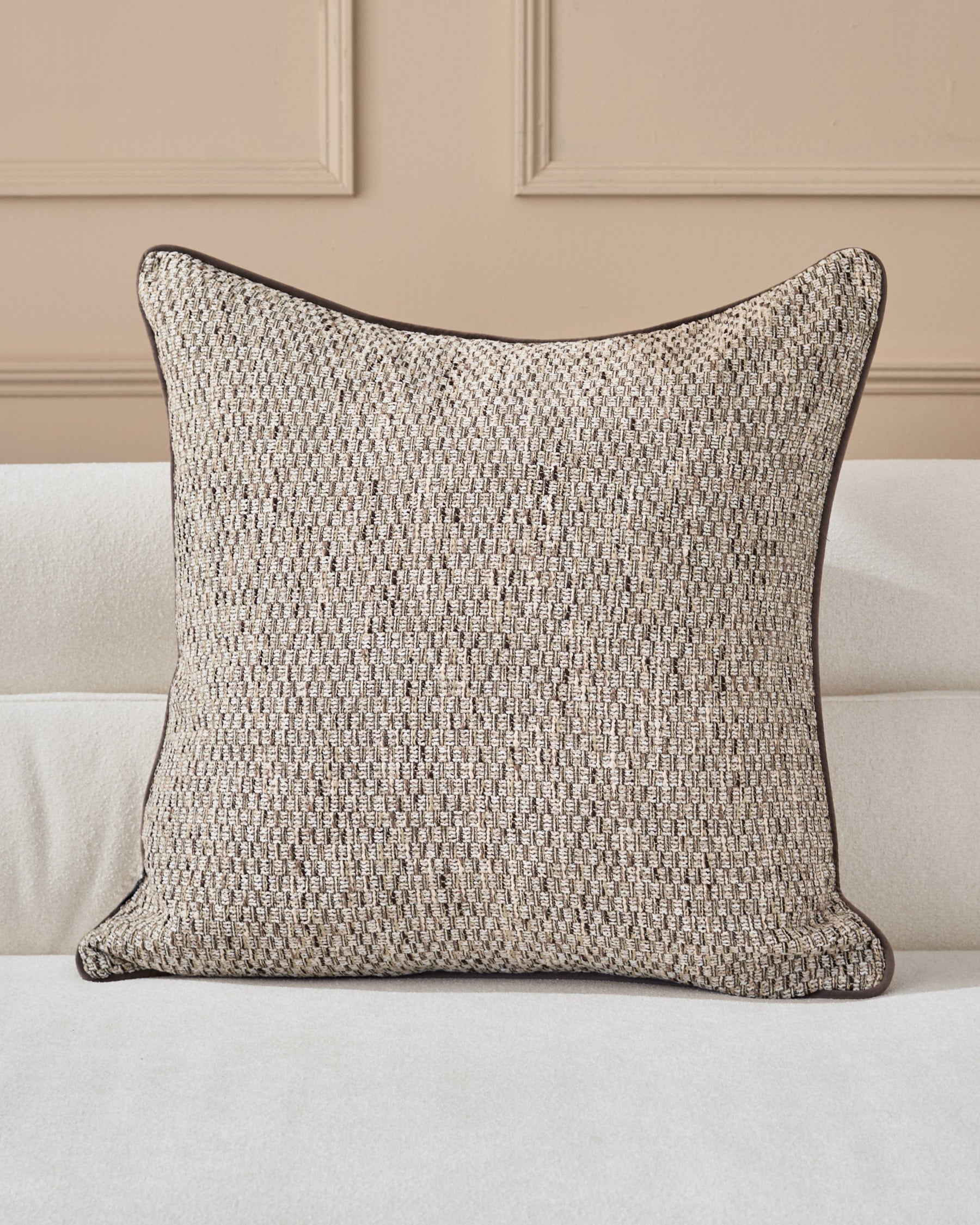 Fabian cushion in natural textured weave with dark piping, displayed on a cream upholstered sofa against a beige paneled wall.