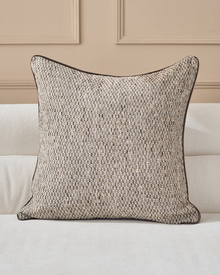 Fabian cushion in natural textured weave with dark piping, displayed on a cream upholstered sofa against a beige paneled wall.
