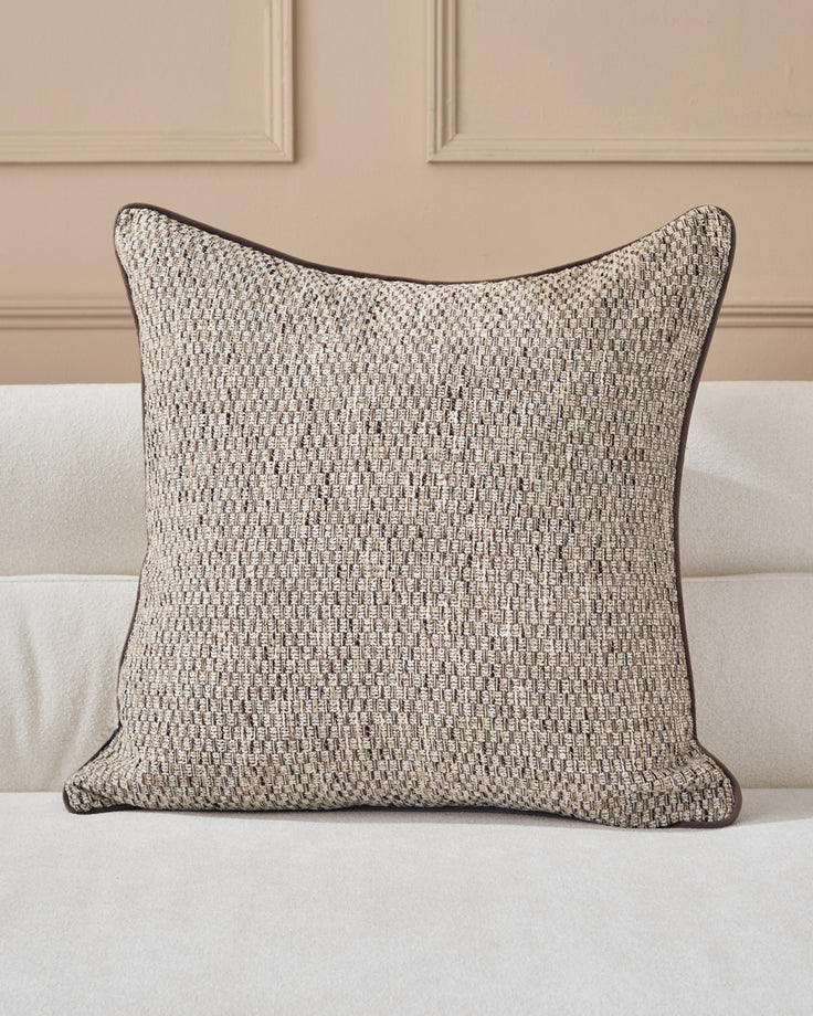 Fabian cushion in natural textured weave with dark piping, displayed on a cream upholstered sofa against a beige paneled wall.