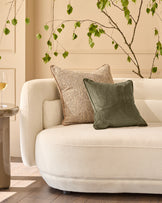 Large green velvet cushion displayed on a cream curved sofa with natural styling and hanging foliage decoration.