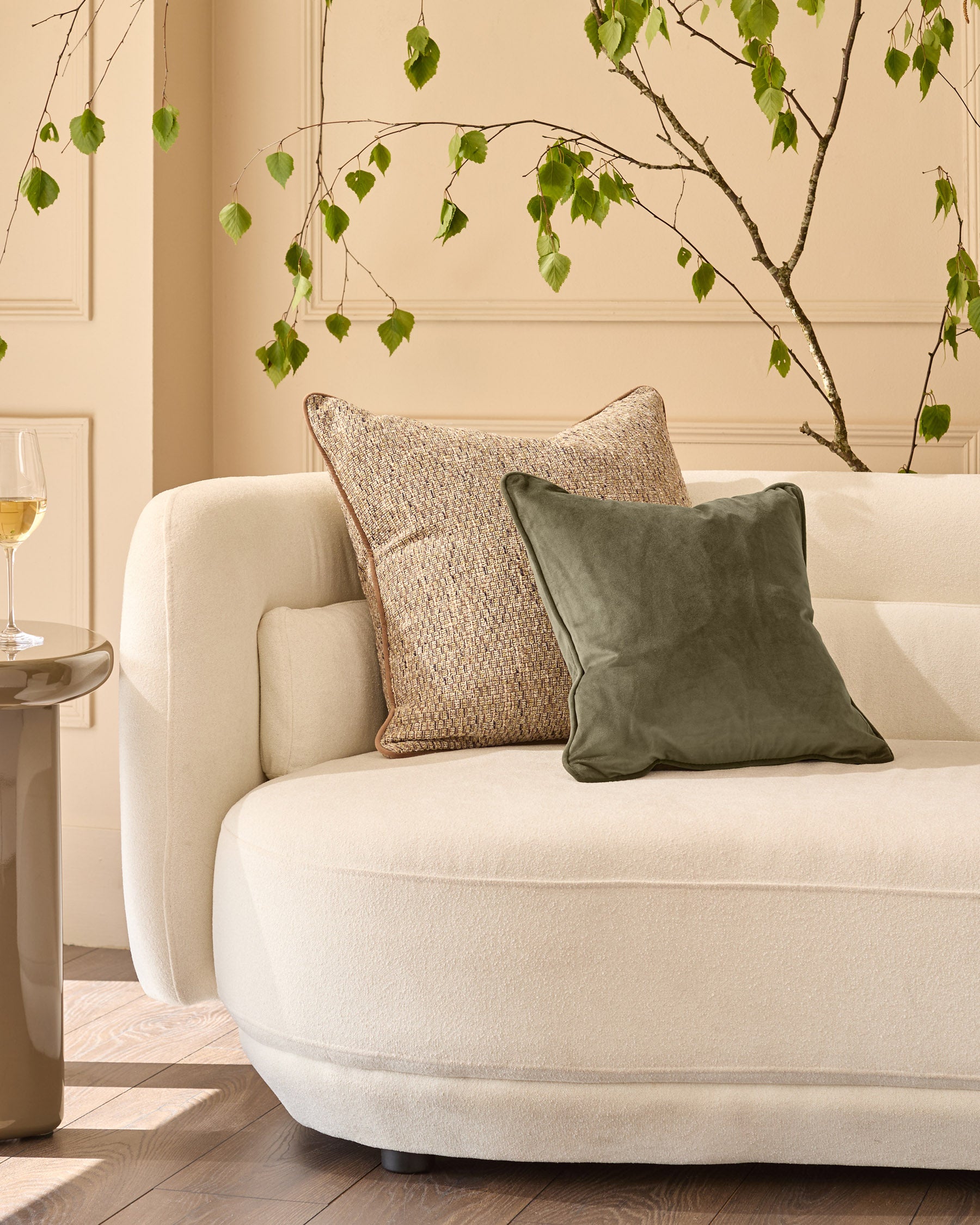Large green velvet cushion displayed on a cream curved sofa with natural styling and hanging foliage decoration.