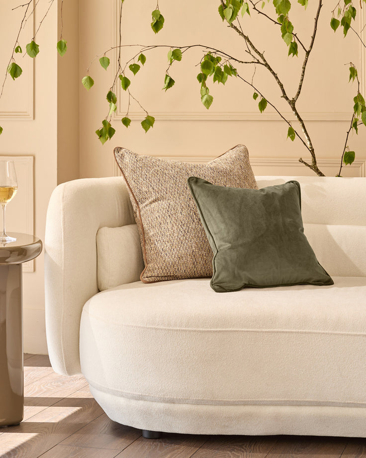 Large green velvet cushion displayed on a cream curved sofa with natural styling and hanging foliage decoration.