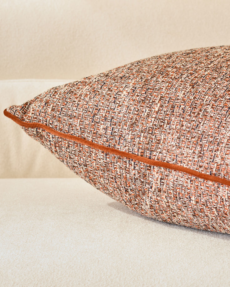 Rust-colored woven lumbar cushion with cream trim, displayed at an angle against a neutral background.