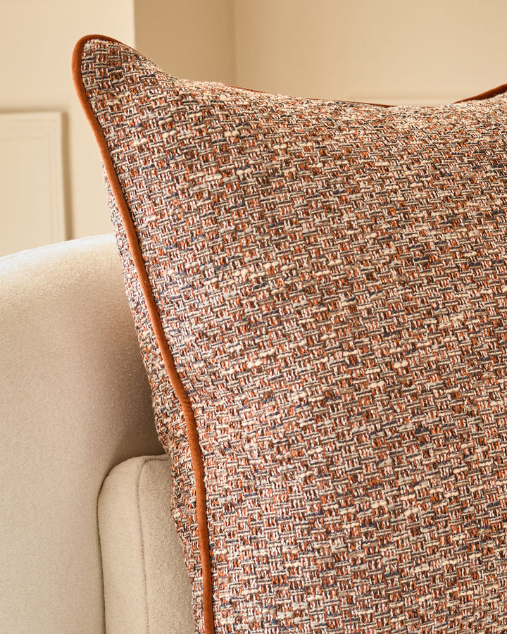 Rust-colored textured cushion with brown leather piping, displayed on a cream upholstered chair.