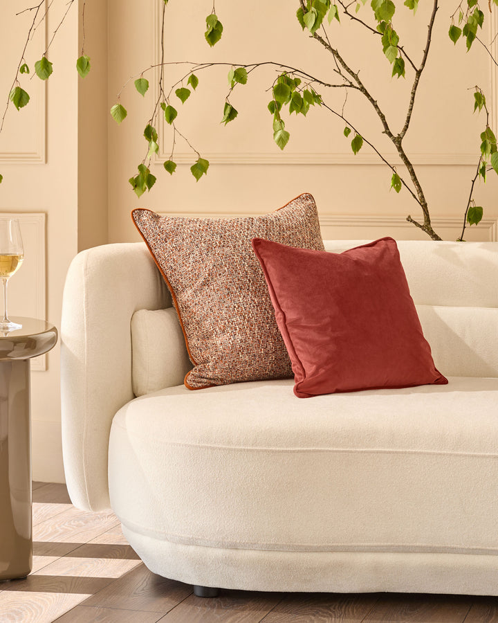 Rust textured cushion with piping displayed on cream curved sofa in bright interior setting.