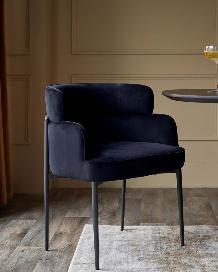 A plush navy velvet chair with a rounded back and sleek, dark metal legs sits on a patterned rug near a small, dark table.