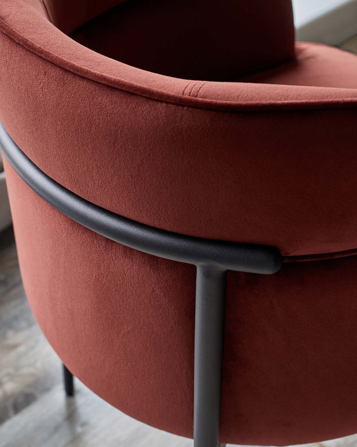 Close-up of a stylish, curved velvet chair in a rich terracotta colour with a sleek, black metal frame.
