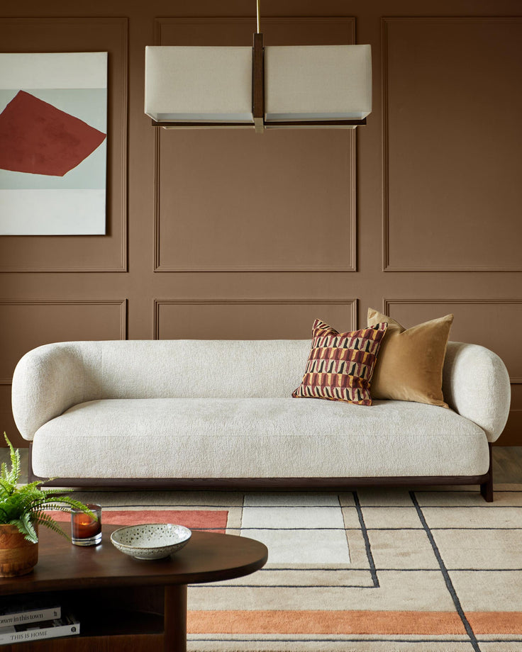 A neutral coloured sofa with brown frame, patterned cushions and a modern light fitting.