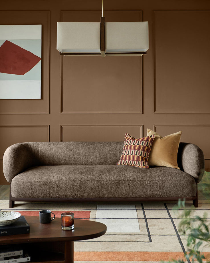 Brown sofa with patterned cushions, under a rectangular pendant light. Coffee table with books, candle & mug in foreground.