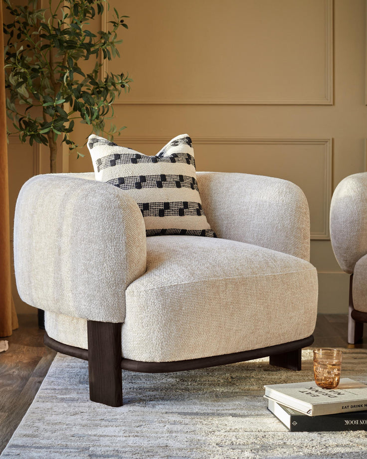 A stylish armchair with a patterned cushion, placed on a neutral rug near a plant and books.