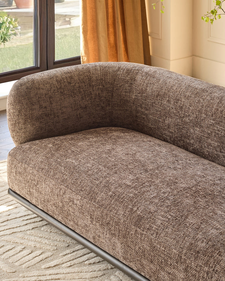 Florence 2-seater sofa in mocha fabric with curved backrest and metal base, shown at an angle in a bright room with windows.