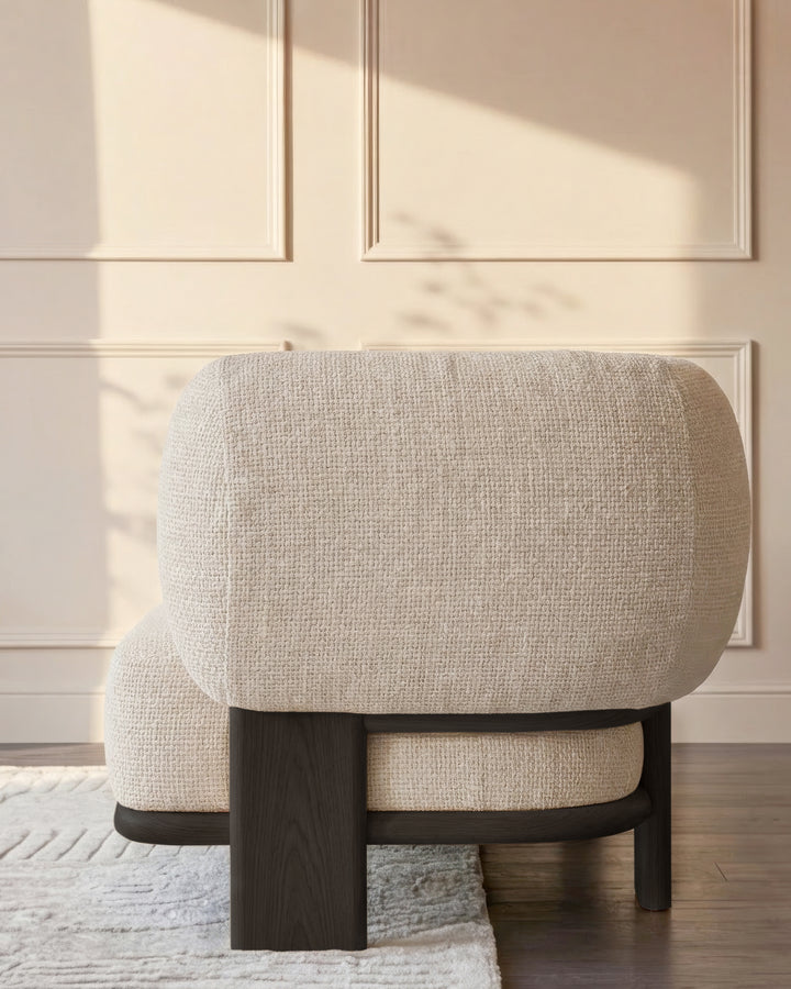 Cream fabric chaise lounge with curved backrest and dark wood frame, shown from rear angle in minimalist room setting.