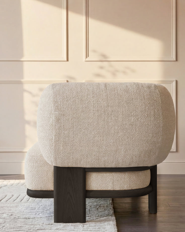 Cream fabric chaise lounge with curved backrest and dark wood frame, shown from rear angle in minimalist room setting.