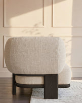 Cream upholstered armchair with curved back and dark wood frame, shown from rear angle in sunlit room.
