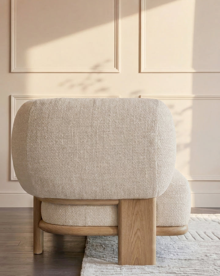 Cream upholstered accent chair with rounded cushioned back and seat, natural wood frame, shown from rear angle in bright sunlit room.