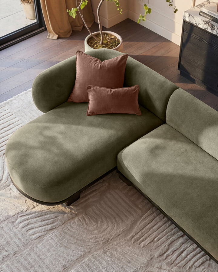 Florence left-hand facing chaise sofa in olive green fabric with dark wood base, styled with burgundy pillows in a modern living room.