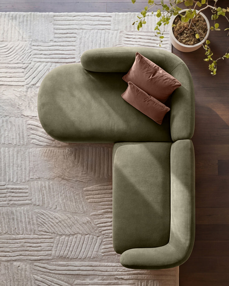 Overhead view of olive green left-hand facing chaise sofa with brown pillows, on textured beige rug with potted plant.