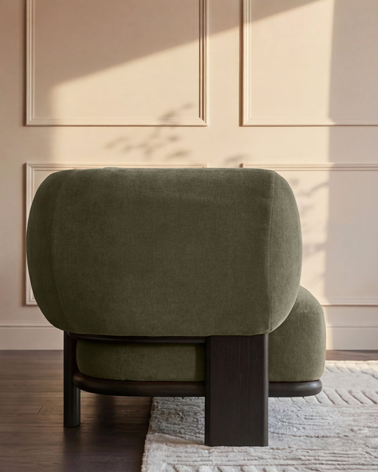 Olive green upholstered armchair with curved back and dark wood base, viewed from behind in a sunlit room.