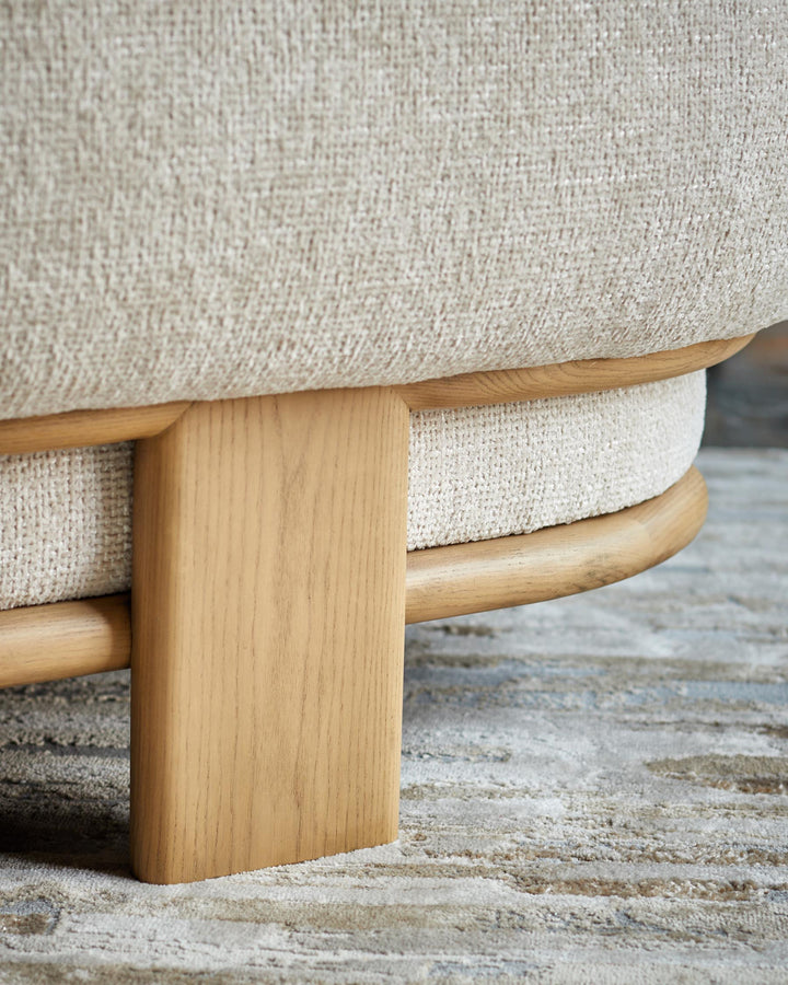 Close-up of a neutral-toned fabric chair with a light wooden frame and leg, sitting on a patterned rug. Modern furniture detail.