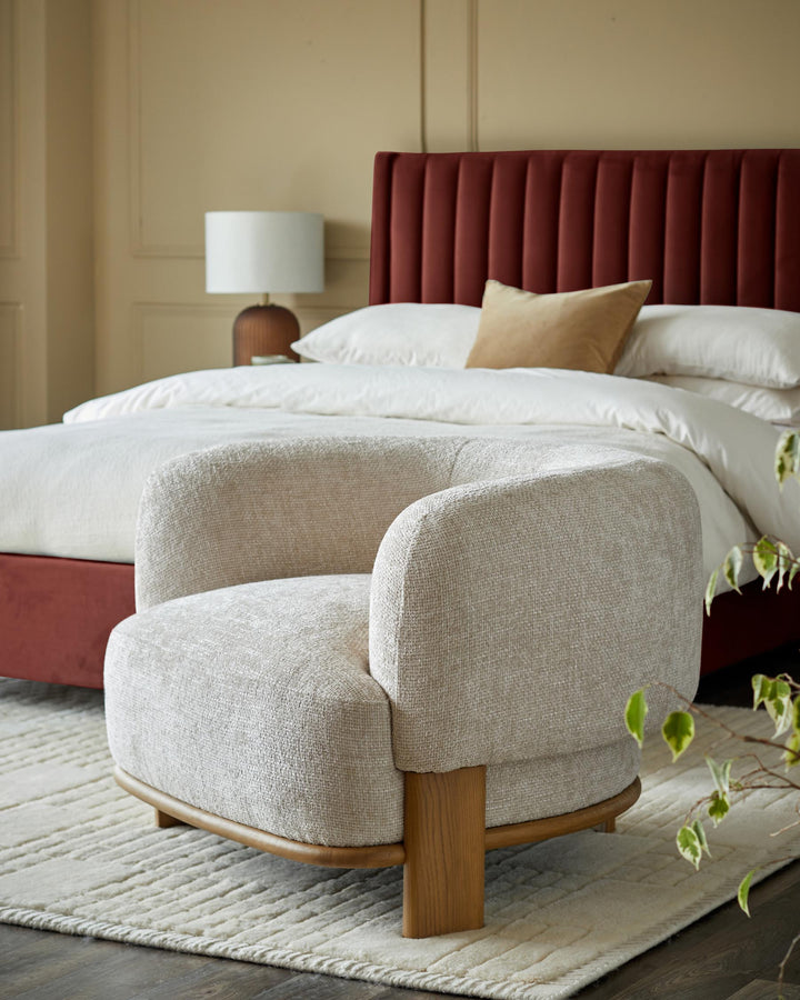 A stylish, textured cream armchair sits on a patterned rug, with a red bedhead and lamp visible in the background.