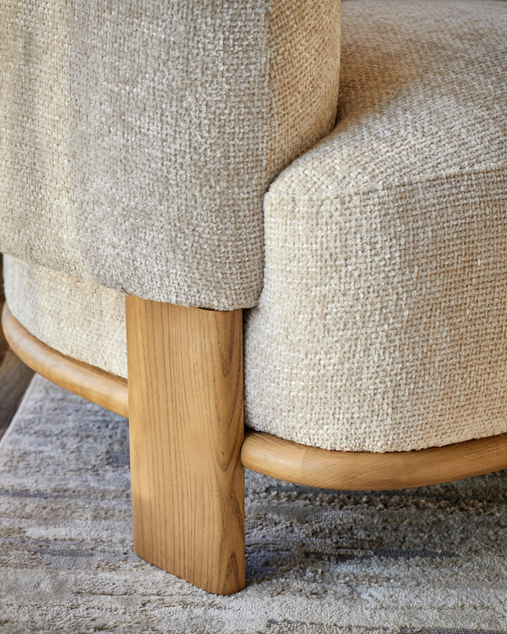 Close-up of an upholstered chair with a light-coloured, textured fabric and wooden legs on a patterned rug.