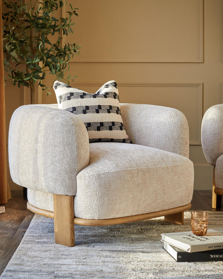 A cosy armchair with a textured fabric, complemented by a patterned cushion and wooden frame. Books and a drink sit nearby.