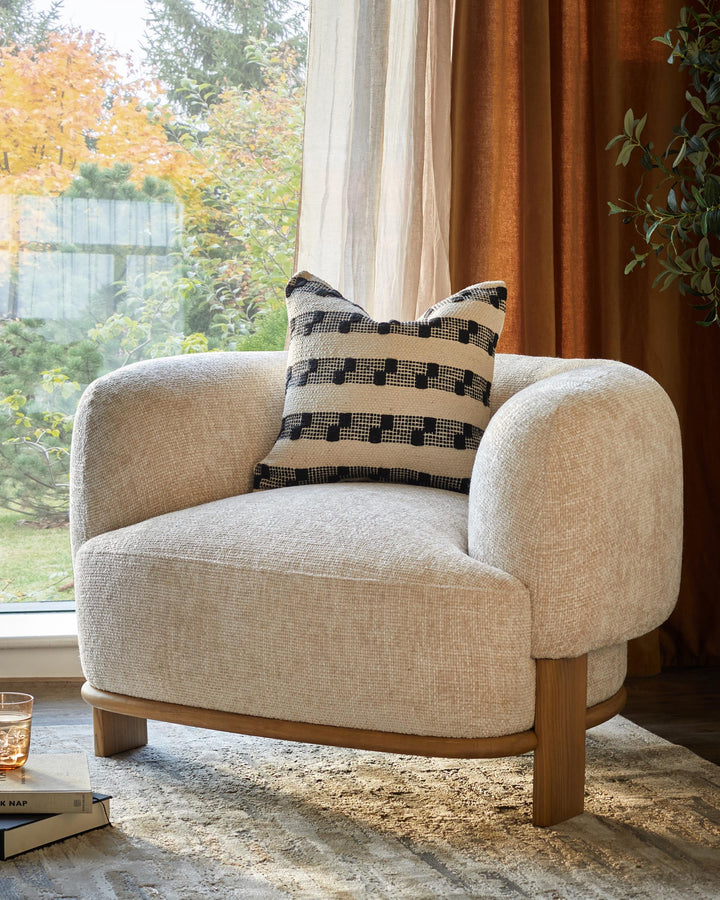 A stylish armchair with a neutral textured fabric, wooden frame and patterned cushion sits near a window.