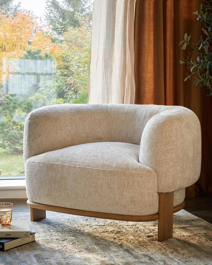 A stylish, comfy, cream armchair with a wooden base sits next to a sunlit window. Perfect for modern living rooms.