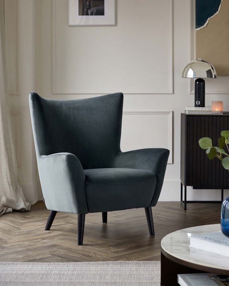 Elegant dark teal wingback chair with velvet upholstery and black wooden legs, positioned next to a modern dark wood side cabinet with a chrome arched table lamp on top, in a chic living space featuring neutral tones.