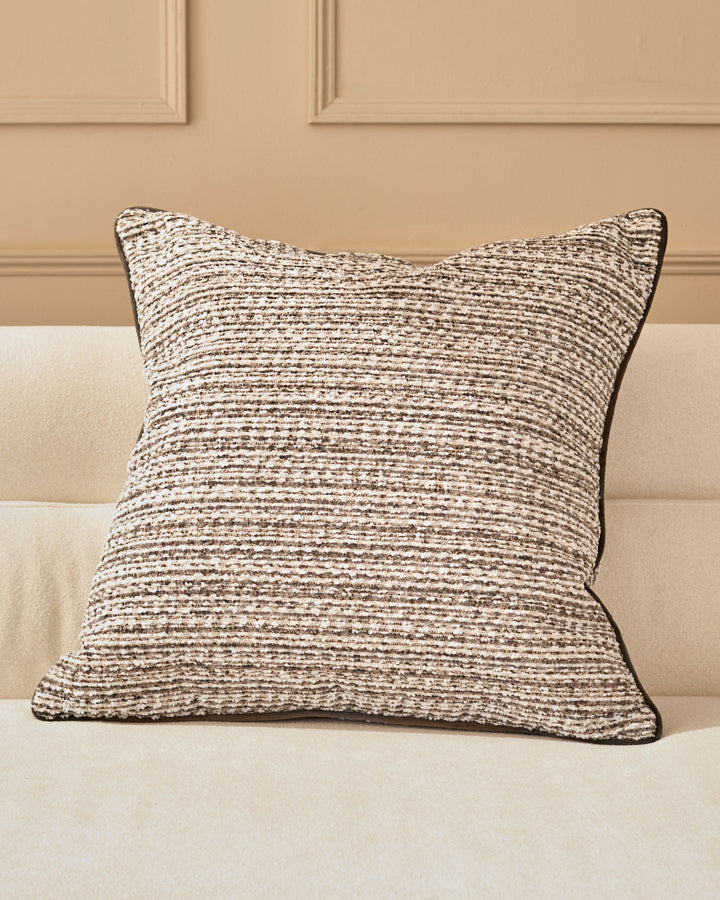 Large Franco cushion in chocolate brown textured fabric with piping detail, displayed on a cream upholstered bed.