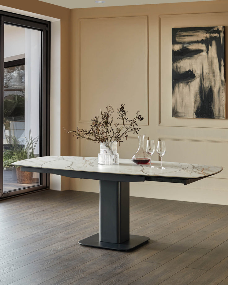 A modern dining table with a marble top and a dark grey pedestal base, complete with a vase and glassware.
