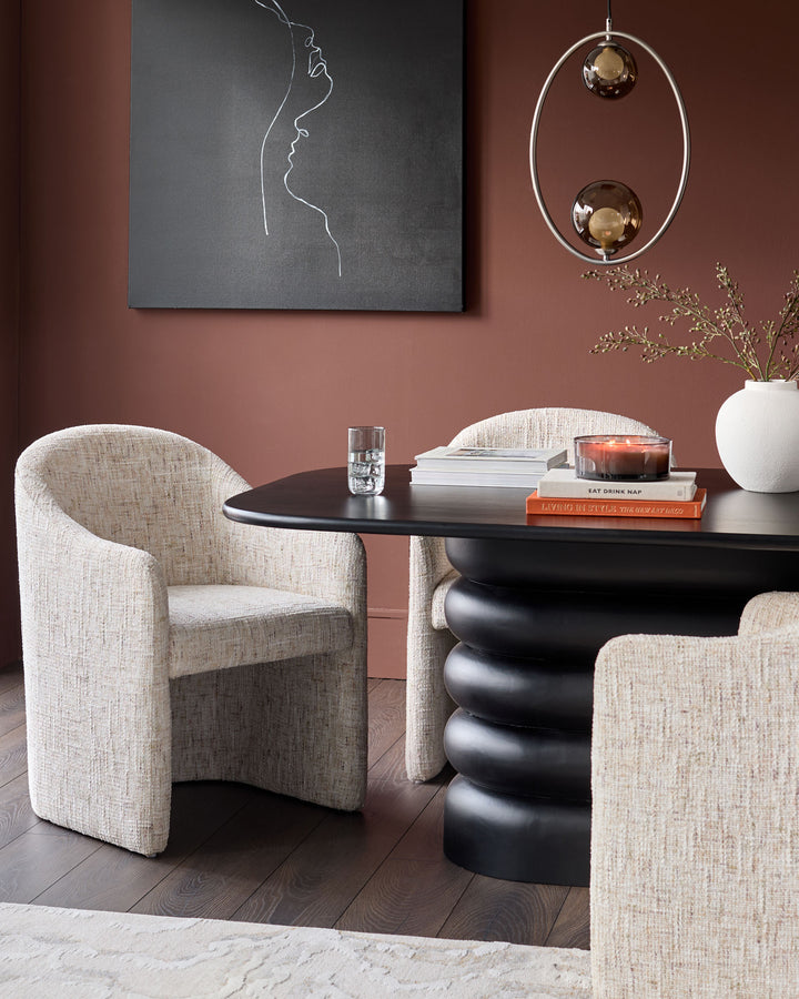 Freya dining chair in natural fabric, shown in a styled room setting with black table and terracotta walls.