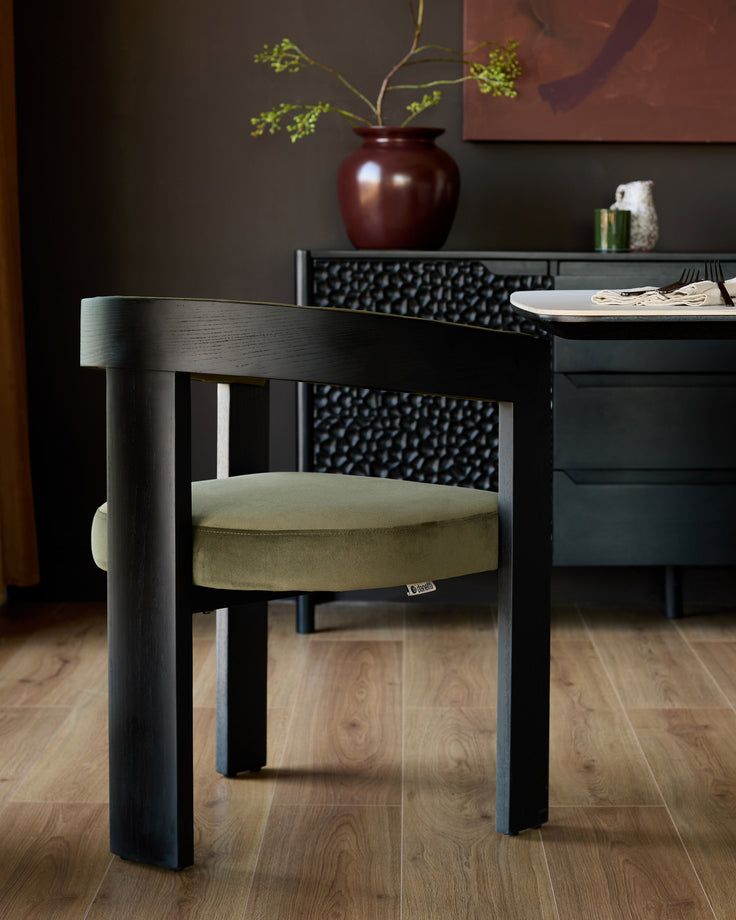 Black wooden dining chair with olive green velvet seat cushion, cylindrical backrest, displayed in modern interior setting.