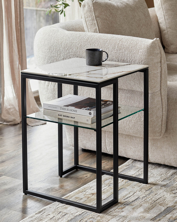 A modern side table with a black frame, marble top and glass shelf, next to a cosy sofa.