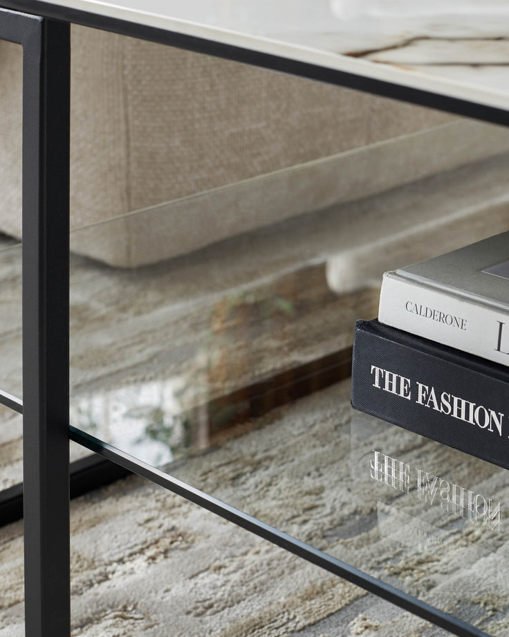 A modern glass coffee table with a black frame. Two books sit on the bottom shelf.