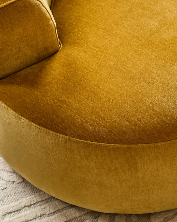 Ochre fabric armchair with curved rounded back and seat, shown from above on a neutral striped rug.