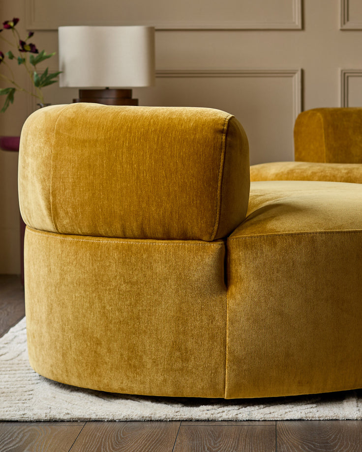 Ochre fabric armchair with curved, rounded silhouette and rolled armrests, shown from side angle in warm, neutral interior.