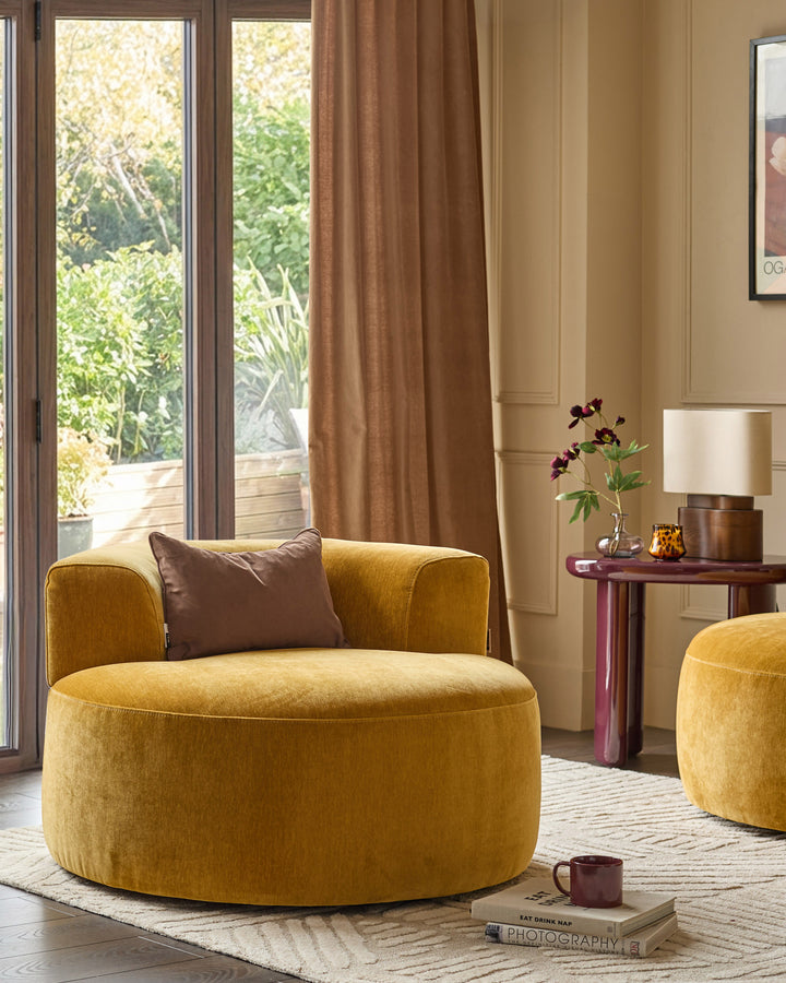 Gianna armchair in ochre fabric with rounded shape, brown accent pillow, shown in warm-toned living room setting.