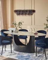 An oval dining table with a marble top and chairs upholstered in navy velvet, illuminated by a modern chandelier.