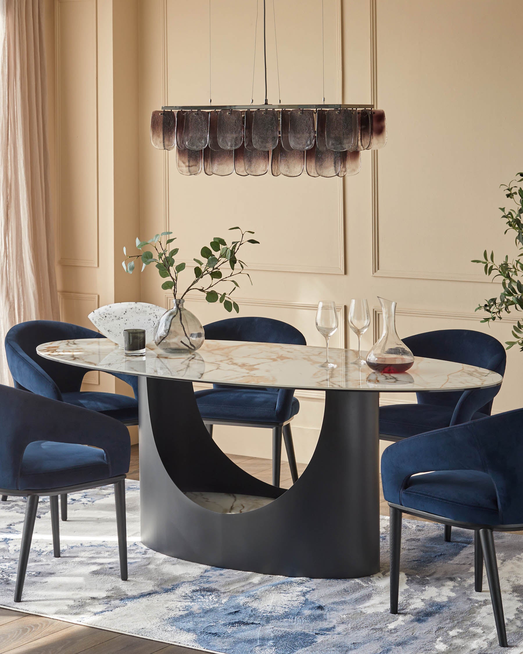An oval dining table with a marble top and chairs upholstered in navy velvet, illuminated by a modern chandelier.